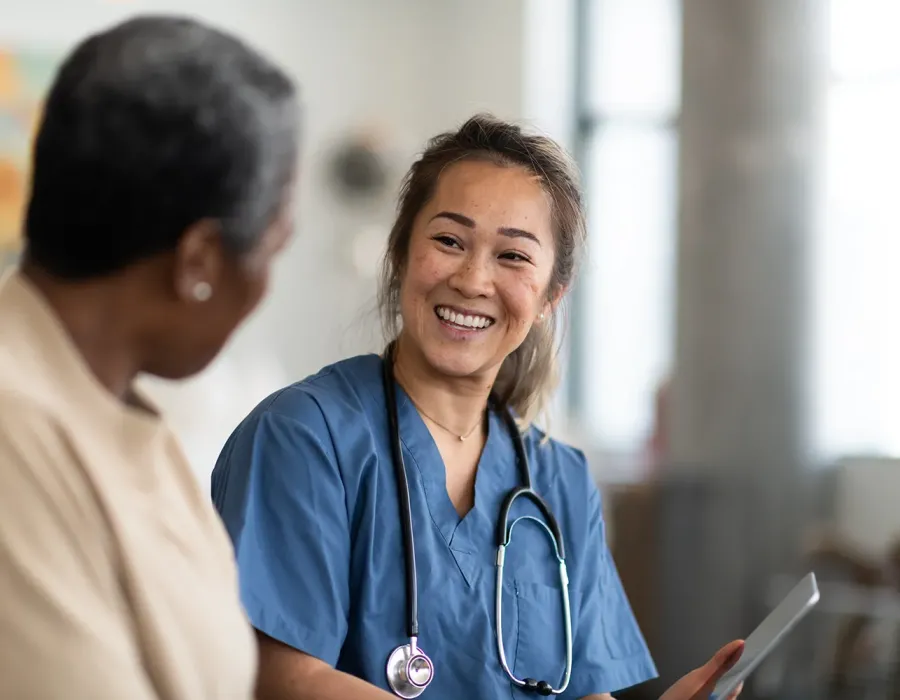 Nurse in blue scrubs with a stethoscope smiles warmly while talking with a patient