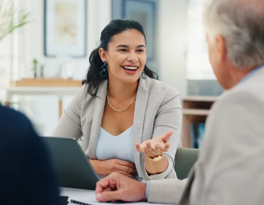 Smiling finance professional speaks with two adults across a table during a meeting, gesturing while discussing information beside an open laptop in a bright office setting.