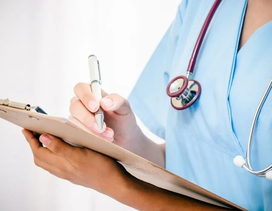 Practical nurse in blue scrubs and wearing stethoscope taking notes on clipboard