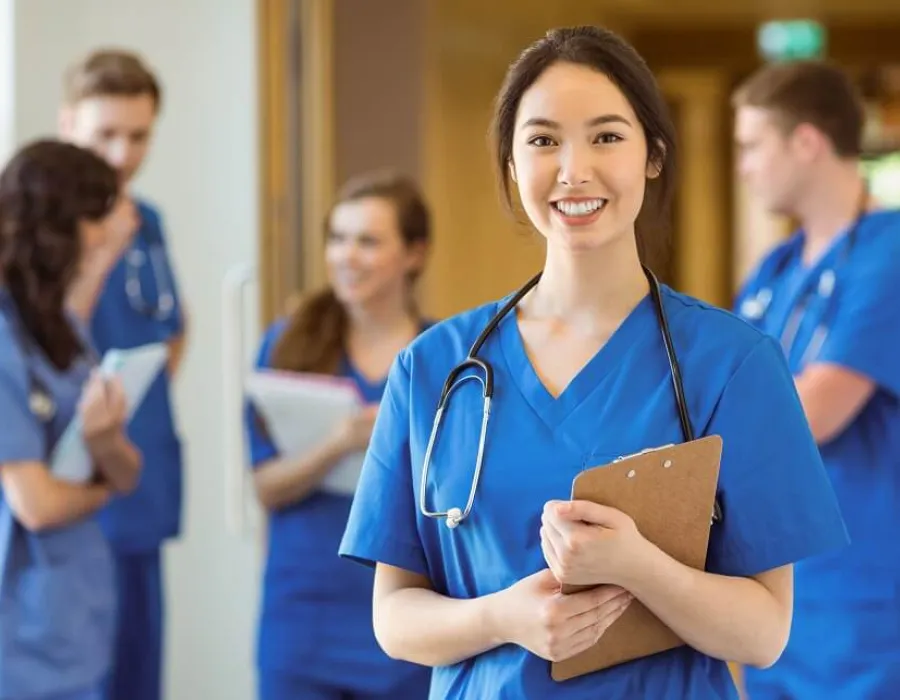 Practical nurse wearing blue scrubs holding clipboard and smiling in hospital corridor