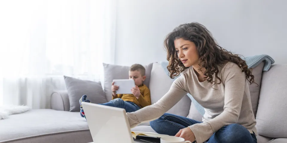 Mother seated on couch taking an online class with son reading on a tablet next to her