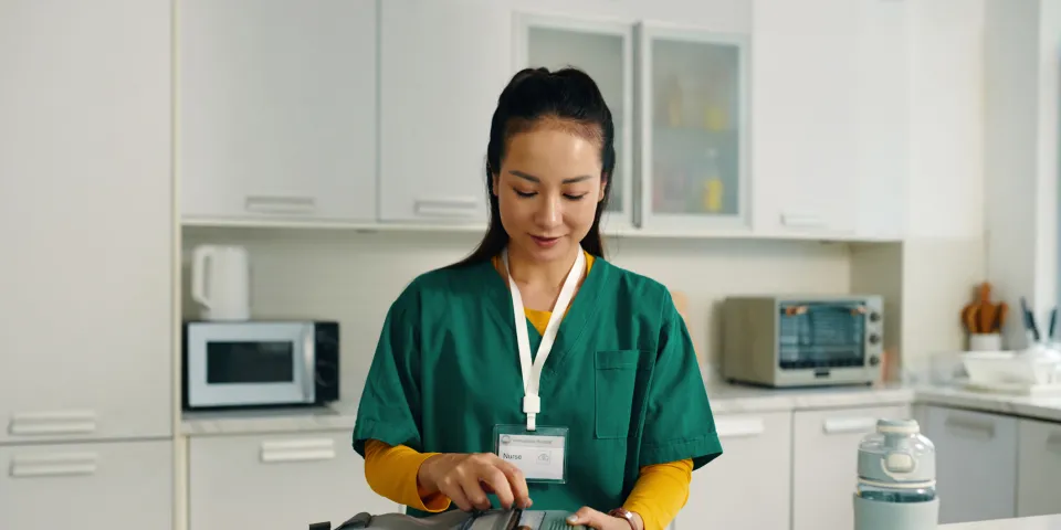 Healthcare professional packing a medical bag while standing in a contemporary kitchen setting with various kitchen appliances visible in background