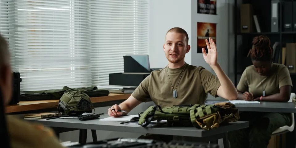 Young male student raising hand and looking at teacher at lesson while asking him question after lecture about military training