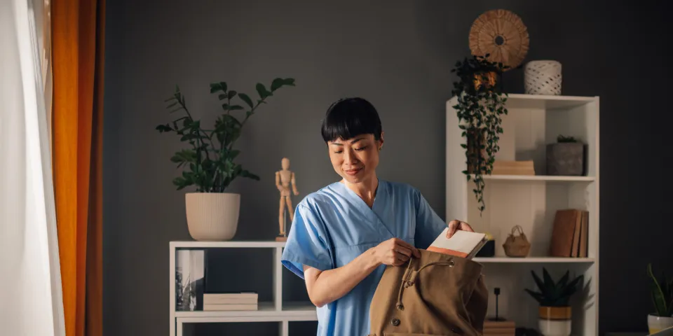 Asian dedicated nurse in blue scrubs is seen preparing medical tools and organizing documents into a brown backpack in a well-lit, modern room with shelves and assorted decor.