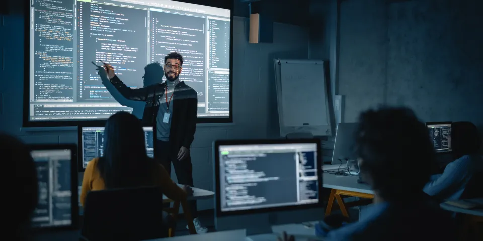 Teacher Giving Computer Science Lecture to Diverse Multiethnic Group of Female and Male Students in Dark College Room. Projecting Slideshow with Programming Code. Explaining Information Technology.