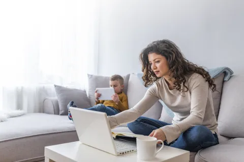 Mother seated on couch taking an online class with son reading on a tablet next to her