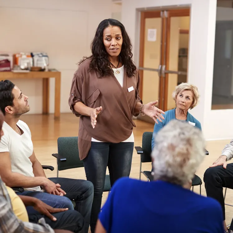 A woman stands and speaks to a diverse group of adults seated in a circle, engaged in a community or support group discussion inside a bright, open room.